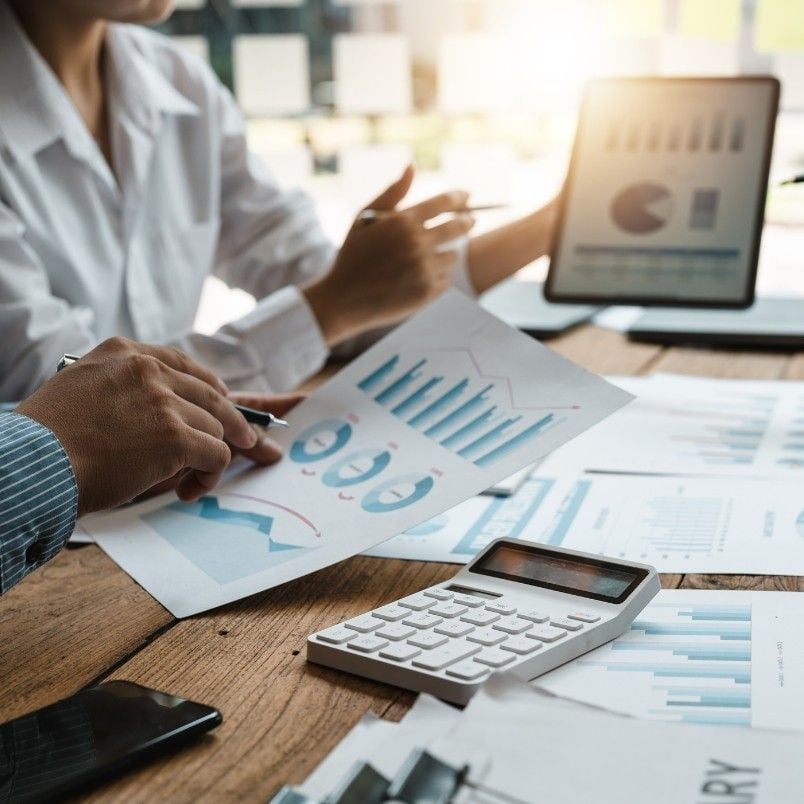 Two people review financial charts and graphs on paper and a tablet at a desk, surrounded by documents and a calculator, discussing how to track project progress as sunlight streams in the background.