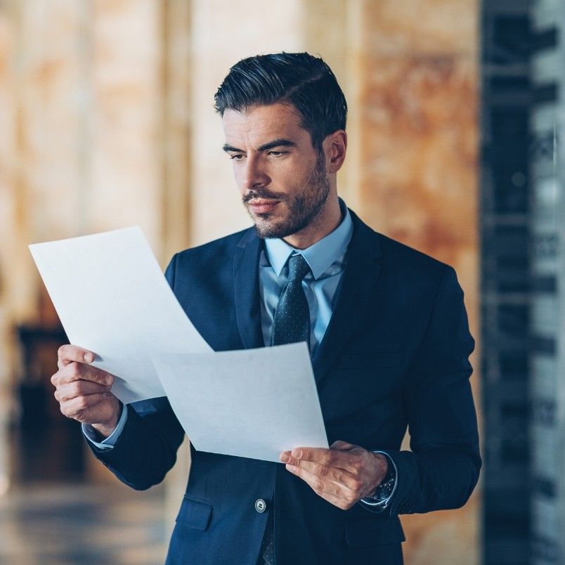 A man in a suit stands indoors, holding and reading two sheets of paper with a focused expression—perhaps reviewing important contract management documents. The background is softly blurred with warm tones.