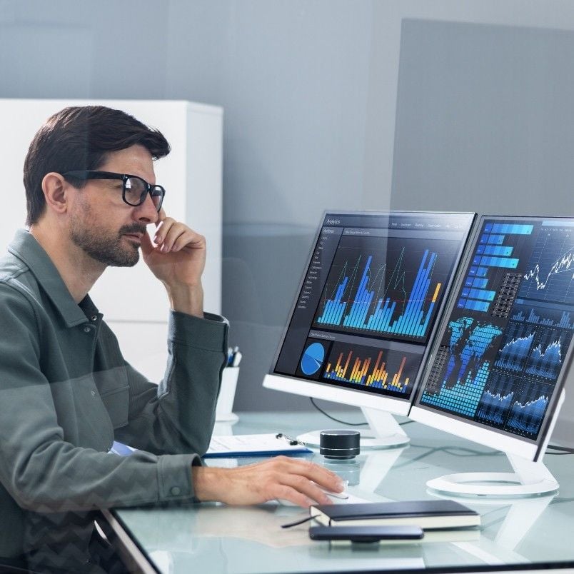 A man wearing glasses sits at a desk, looking thoughtfully at two computer monitors—one displaying colorful data charts and graphs, the other showcasing a wireframe template—in a modern office setting.