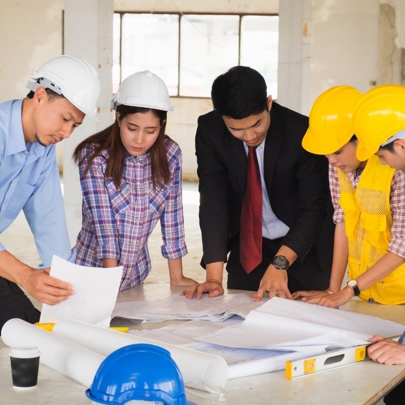 A group of five people, including engineers in hard hats and a man in a suit, gather around a table reviewing blueprints and documents, discussing Construction Project Management at a construction site.