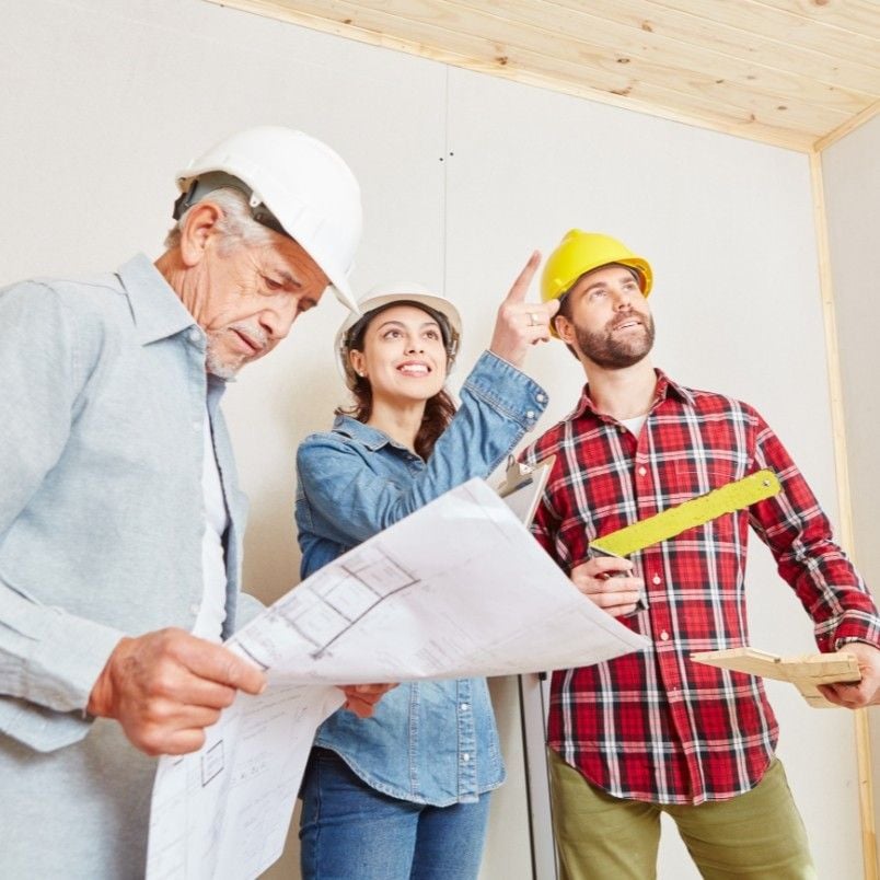 Three construction workers wearing hard hats are inspecting a room. One man holds blueprints, the woman is smiling and pointing upward, and another man in a yellow hard hat, focused on Construction Project Management, holds a measuring tool and wooden planks.