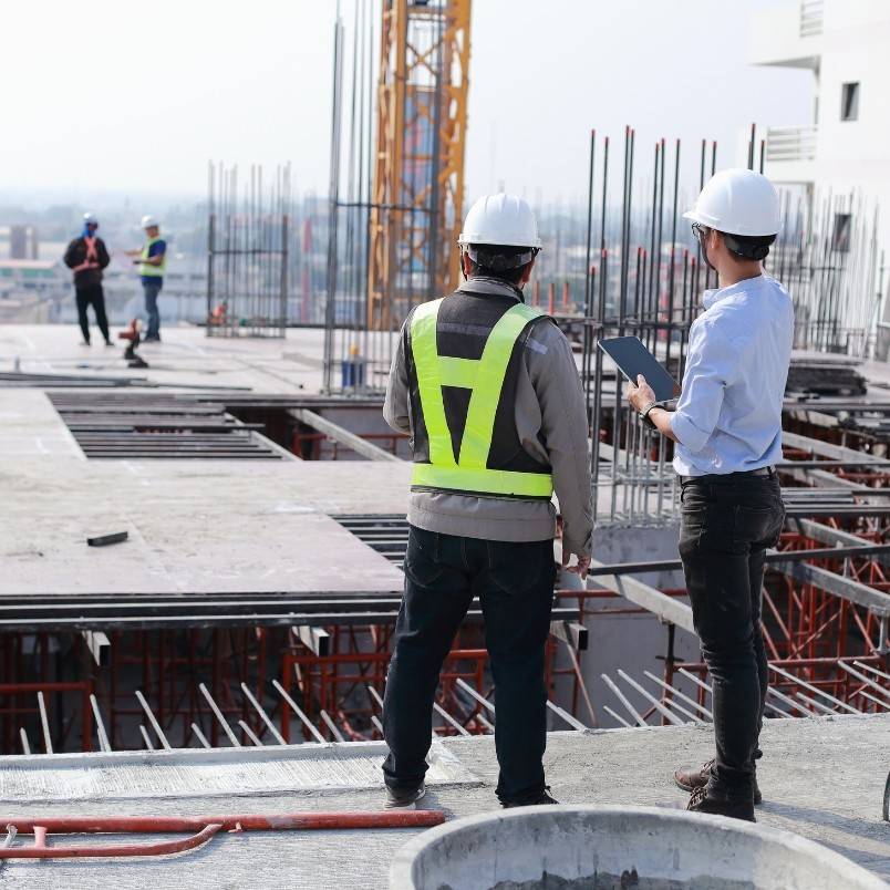 Two construction workers in safety gear and hard hats stand on a building site, overseeing progress and practicing effective Construction Project Management. Rebar, concrete slabs, more workers, and a crane are visible in the background.