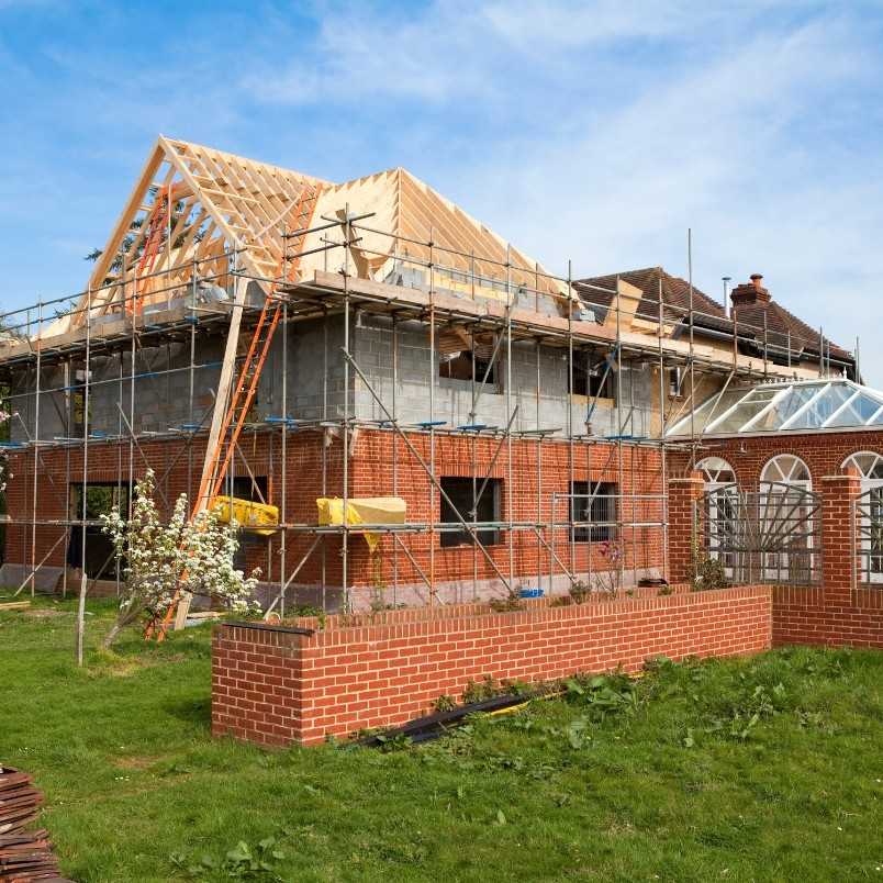 A house under renovation showcases construction project management with scaffolding, exposed roof framing, and building materials. The lower part features red brick walls, while a grassy lawn stretches across the foreground.