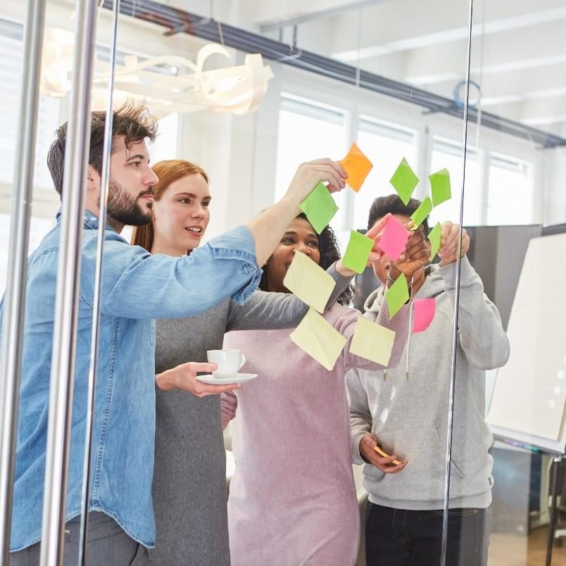 Four people stand together in an office, brainstorming as they place colorful sticky notes on a glass wall. One person holds a cup and saucer while the group collaborates and discusses ideas in a bright, modern workspace.