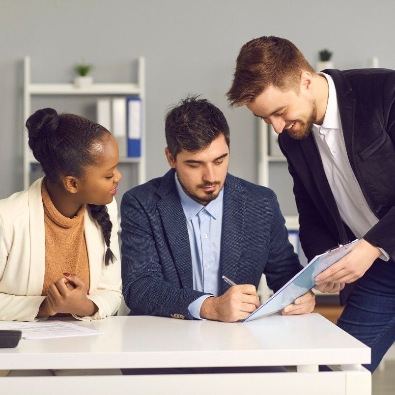 Three colleagues in business attire discuss documents at a desk, focusing on contract management. One man is seated and writing, while a woman and another man stand beside him, reviewing papers together in an office setting.