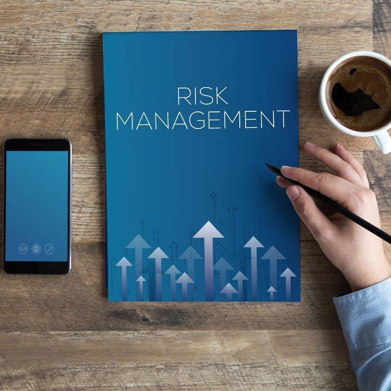 A person’s hand holding a pen next to a blue "Risk Management" folder with upward arrows sits on a wooden desk, alongside a risk register, smartphone, and a cup of coffee.