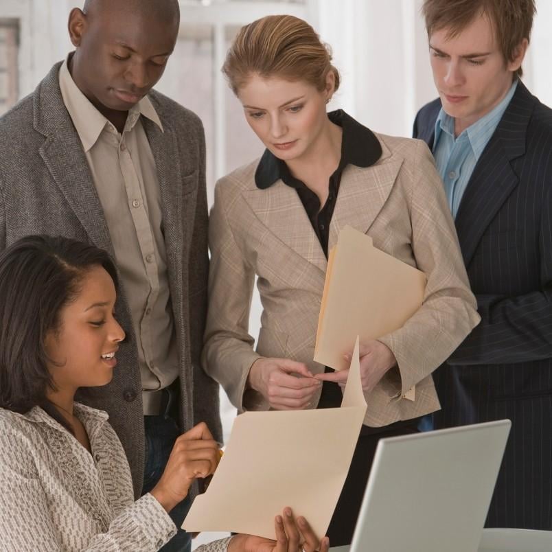 Four professionally dressed colleagues gather around a desk, reviewing documents together. One woman points at a file detailing the project communication plan, while the others stand attentively, suggesting a collaborative work discussion.