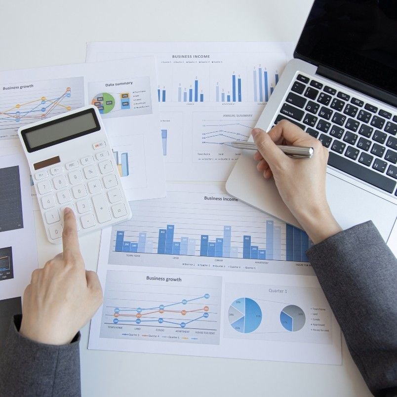 Person analyzing financial charts and graphs on printed papers, using a calculator and writing with a pen, with an open laptop nearby. The workspace suggests financial planning or alternative analysis activities.