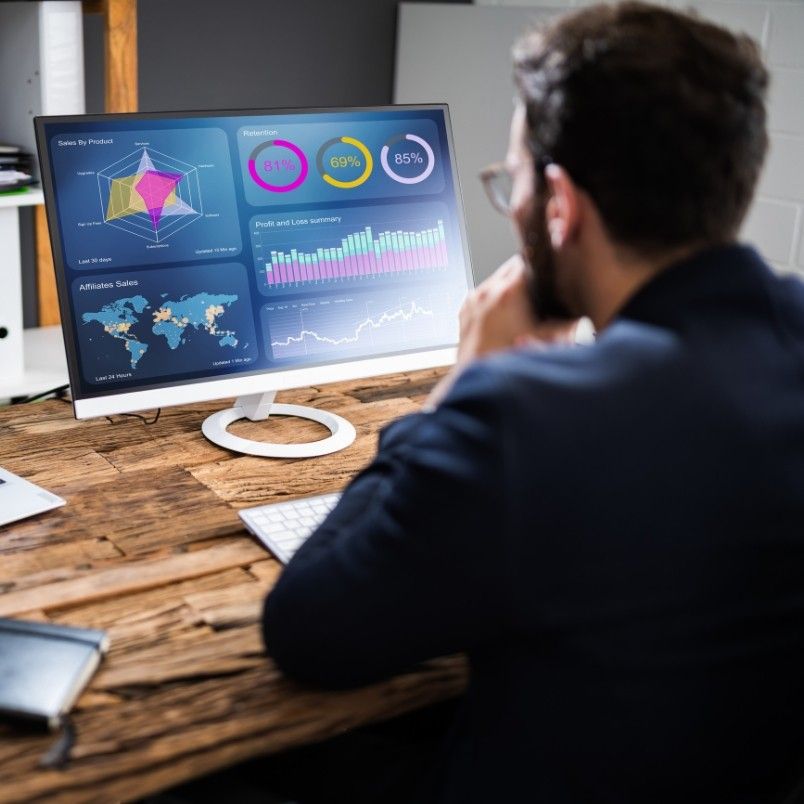 A person in a suit sits at a wooden desk, analyzing colorful charts, graphs, and maps related to project management workflow displayed on a computer monitor in an office setting.
