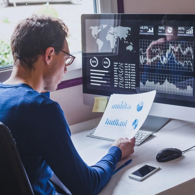 A person sits at a desk analyzing printed charts on how to track project progress, with a computer displaying various global data visualizations and graphs. A smartphone, mouse, and pen are also on the desk.