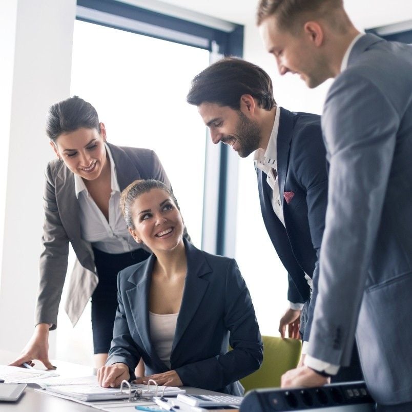 Four business professionals in formal attire gather around a desk in a bright office, smiling and engaging in a collaborative discussion focused on project management and business process improvement. One woman is seated while the others stand around her.