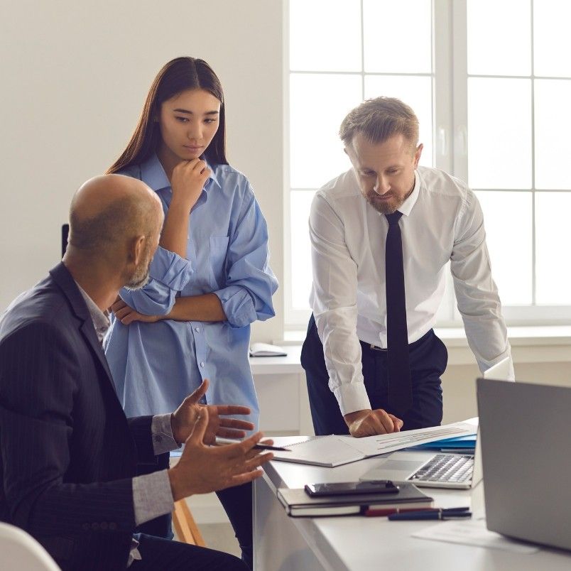 Three people in an office discuss a project management workflow around a desk with documents and laptops. One man gestures while talking, a woman listens thoughtfully, and another man leans forward, focused on the papers.