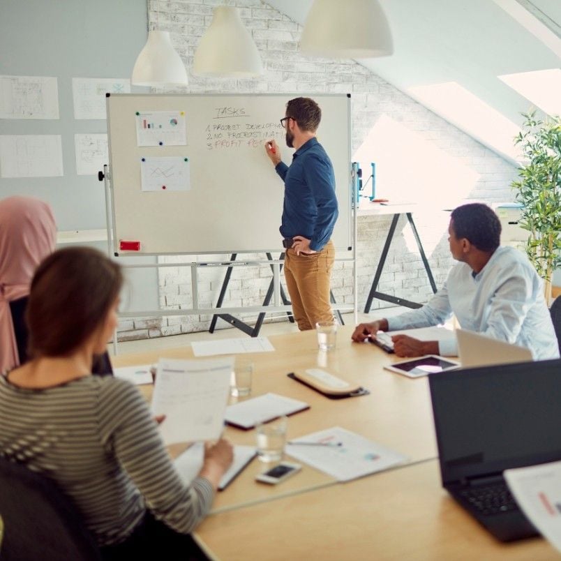 A man demonstrates the project manager role in Scrum at a whiteboard, presenting charts and notes to four colleagues seated around a table in a modern office with laptops, papers, and glasses of water.