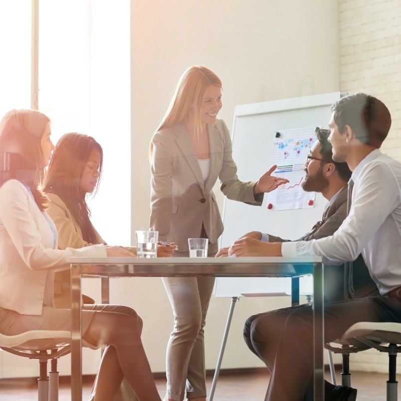 Four people sit at a table while a woman stands, smiling and leading a discussion on project management. A flip chart with business process diagrams is in the background, as sunlight fills the modern office space.