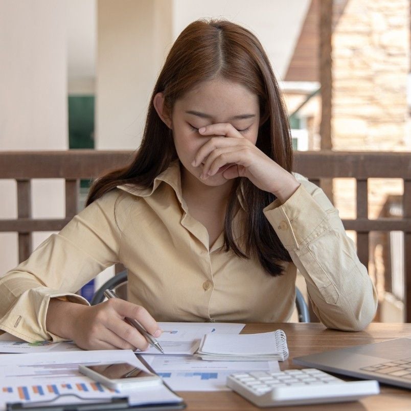 A woman sits at a desk with papers, a calculator, and graphs, looking stressed as she pinches the bridge of her nose with her eyes closed next to a wireframe template.