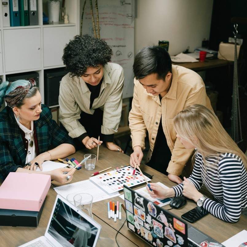 Four people gather around a table with color samples, pens, and laptops, collaboratively working on a creative project and discussing a story map template in a modern office setting.