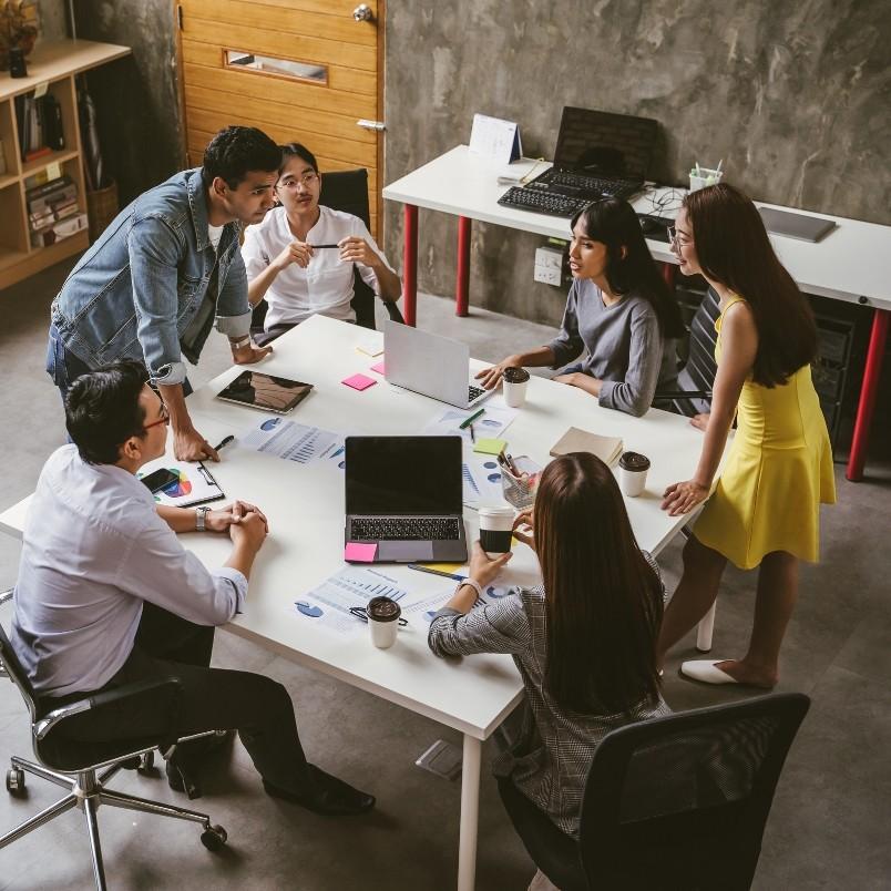 Six people sit and stand around a table in an office, engaged in a collaborative project management workflow. Laptops, documents, and coffee cups are on the table, reflecting a focused and productive atmosphere.