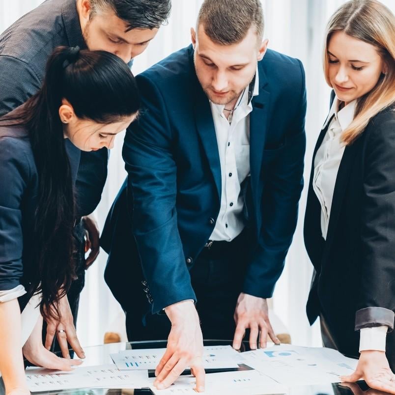 Four business professionals stand around a table, closely examining and discussing documents and charts in front of them—an agile project management example during a collaborative meeting.