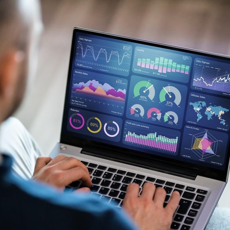 A person using a laptop displaying colorful data dashboards with charts, graphs, and statistics, set within a modern wireframe template, including line graphs, pie charts, bar graphs, and a world map representing data analytics.