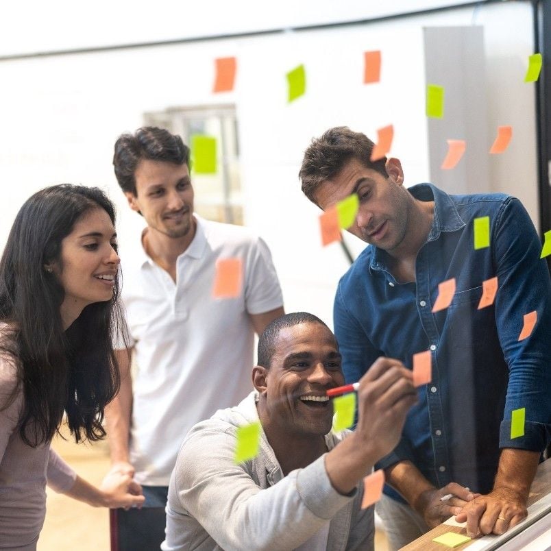Four people brainstorming together, smiling and placing colorful sticky notes on a glass wall in a bright, modern office setting, showcasing their agile skills.
