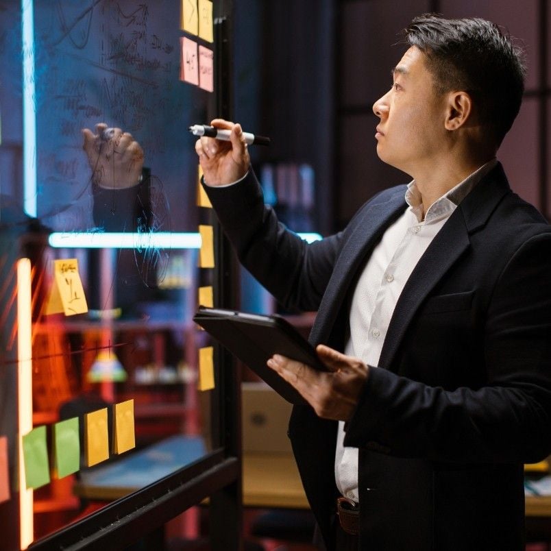 A man in a suit writes on a glass board with a marker while holding a tablet, outlining an agile project charter. Colorful sticky notes cover the glass, and bookshelves are visible in the background of a modern office.