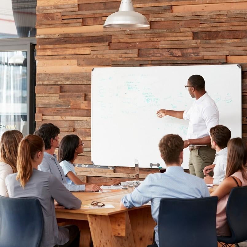 A man stands and points at a whiteboard covered in notes, presenting the project manager role in Scrum to a group of six people seated at a table in a modern office with a wooden accent wall.