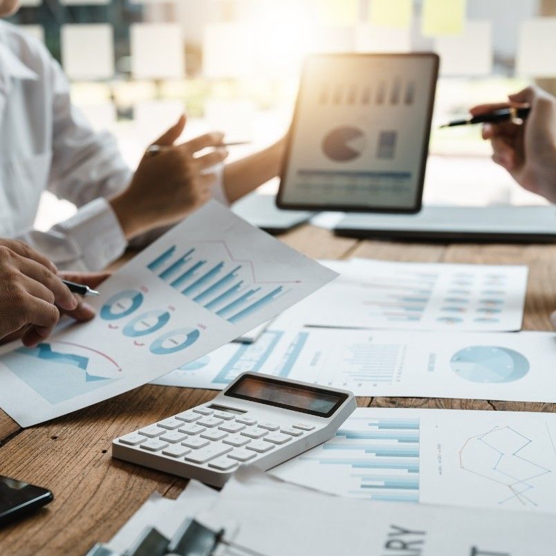 Close-up of people conducting root cause analysis while reviewing financial charts and graphs on printed papers and a tablet at a wooden desk, with a calculator and pens visible, in a bright office setting.