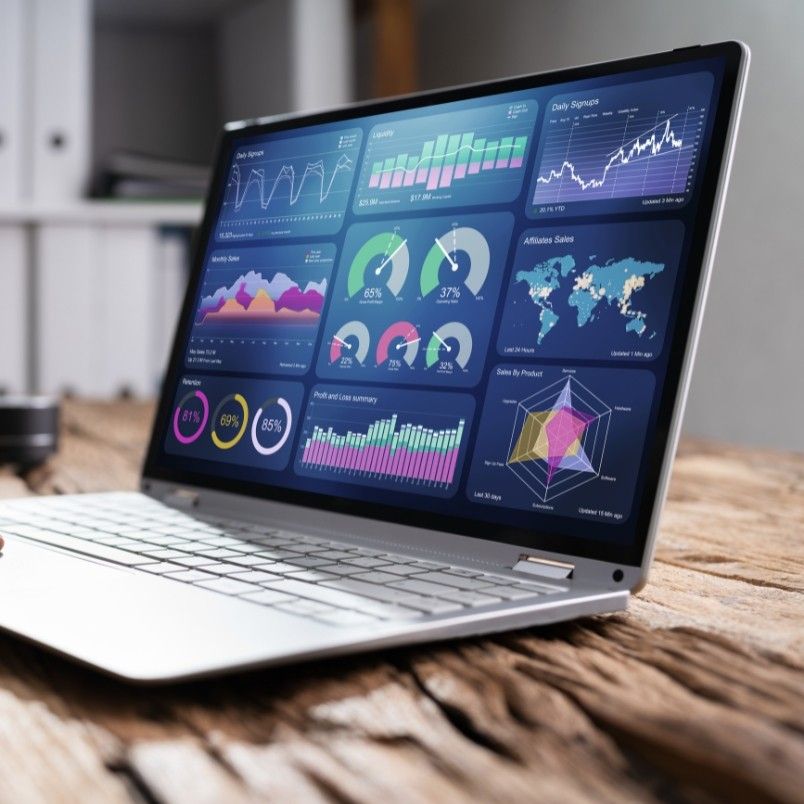 A laptop on a wooden desk displays colorful data analytics dashboards with charts, graphs, and a world map, visualizing business performance metrics and statistics within an organized project management workflow.