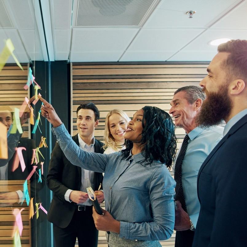 Five professionals stand by a glass wall covered with colorful sticky notes. A woman in front points at a note, while the group smiles and watches, engaged in an Agile Team Charter discussion or brainstorming session.
