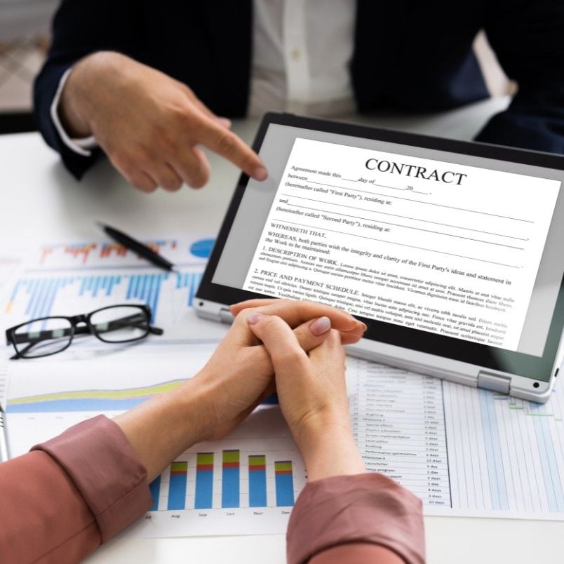 Two people sit at a desk; one points to a laptop displaying a contract, highlighting effective contract management, while the other has hands clasped. Charts and graphs with financial data are spread across the desk.