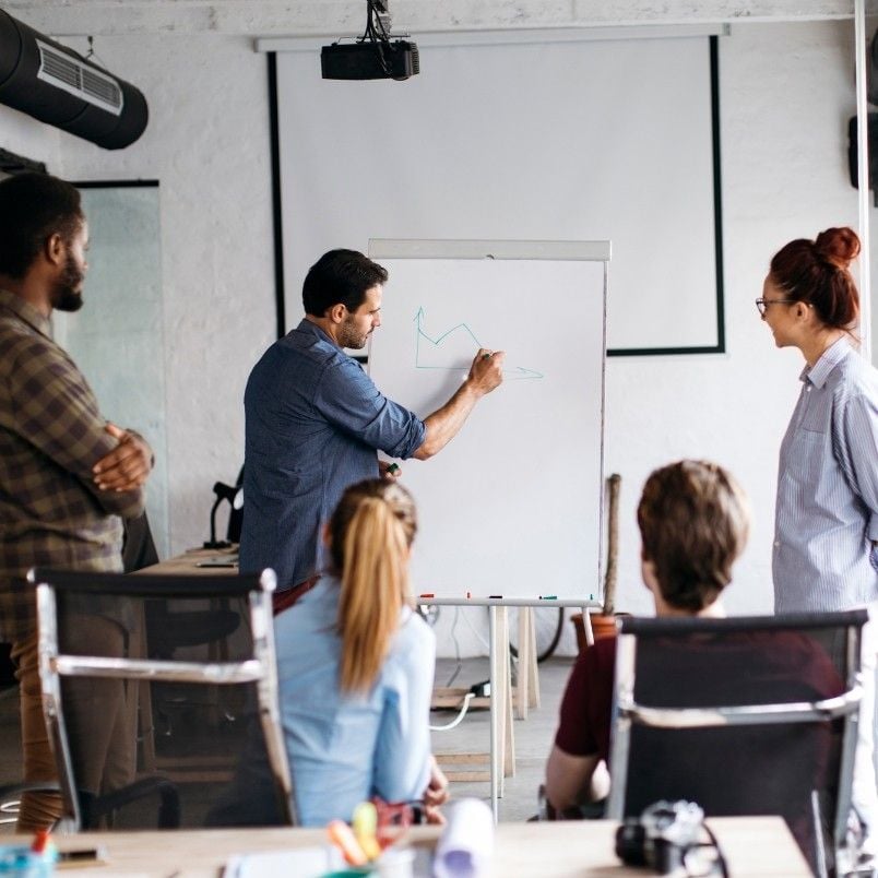 A man explains the project manager role in scrum by drawing a graph on a flip chart while four colleagues watch and listen in a modern office meeting room with chairs and a projector.