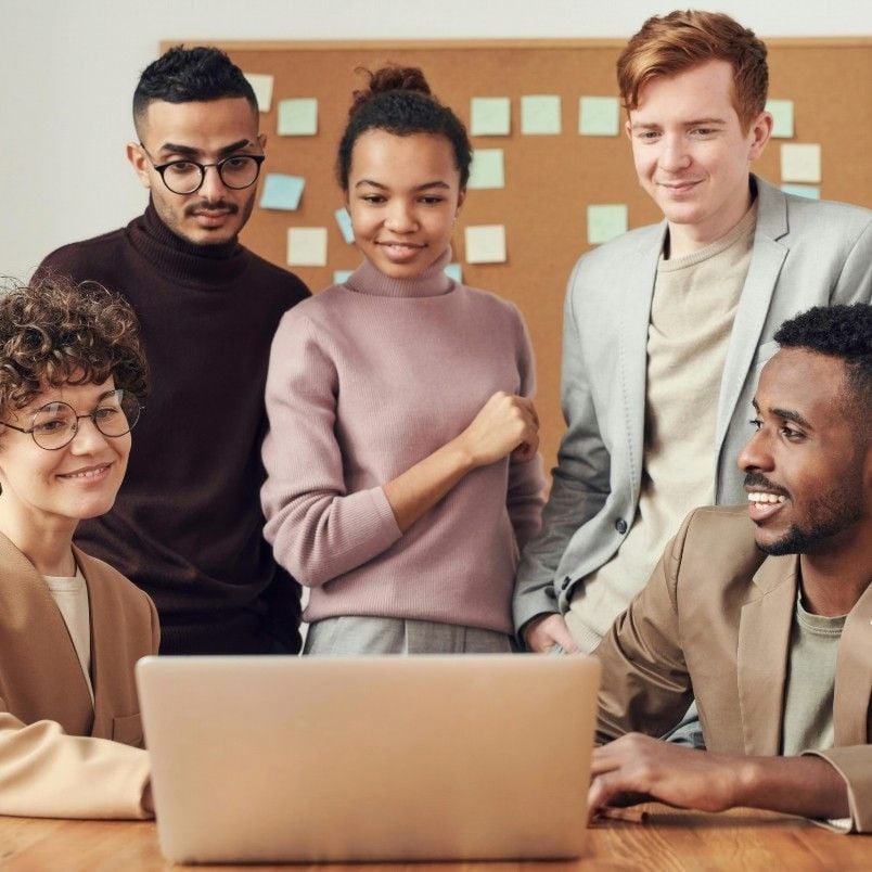 Five people gather around a laptop, smiling and collaborating in an office setting as they discuss their Agile Team Charter. A corkboard with colorful sticky notes is visible in the background.