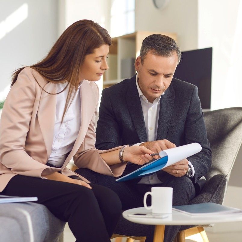 Two business professionals sit together in an office, reviewing contract management documents on a clipboard. The woman points at the papers while the man listens attentively. A coffee cup and laptop are on the table in front of them.