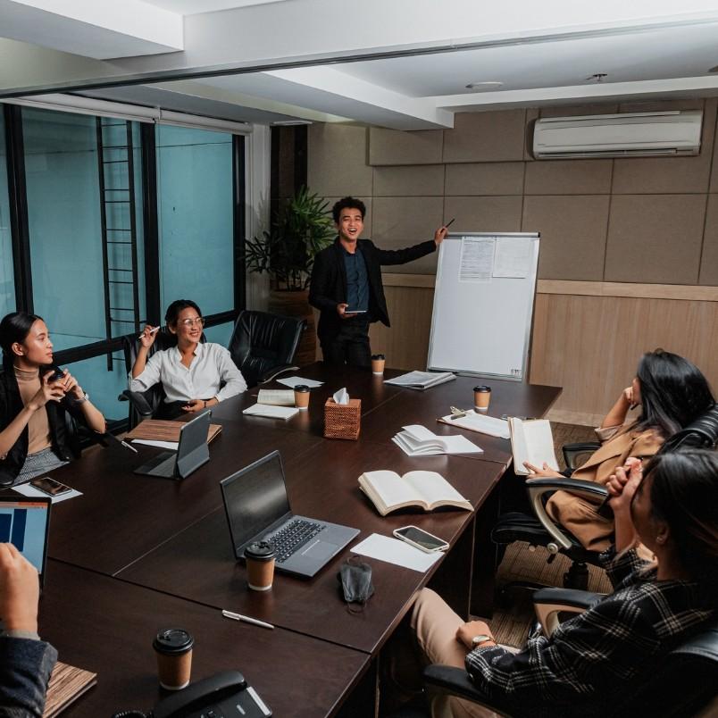 A group of six people sit around a conference table with laptops and coffee cups as one person presents an Agile Team Charter using a whiteboard in a modern office setting.
