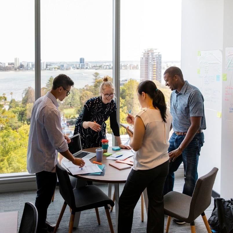 Four people stand around a small round table in a modern office, discussing a project communication plan and reviewing documents on a laptop, with large windows offering views of the cityscape and river in the background.