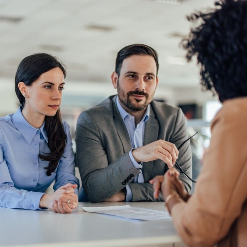Two professionals, a woman in a blue blouse and a man in a gray suit, sit at a table listening attentively to another woman with curly hair discussing contract management in a modern office setting.