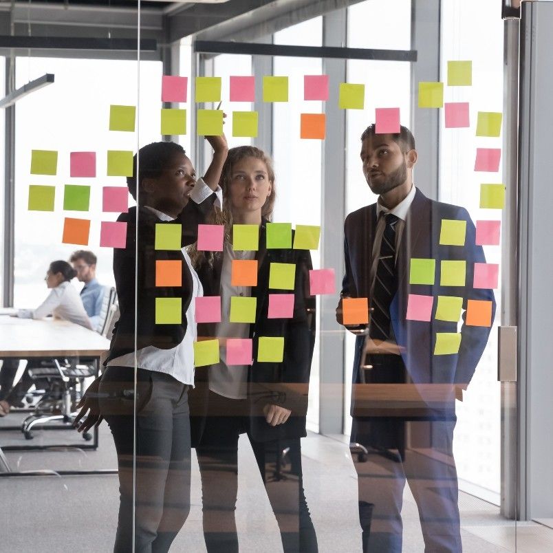 Three business professionals stand in front of a glass wall covered with colorful sticky notes, collaborating on their Agile Team Charter. An office setting with colleagues working in the background is visible.
