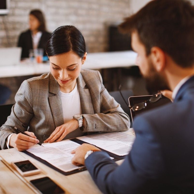A woman in a blazer signs a document on a clipboard, focused on contract management, while a man in a suit sits across from her and points at the paperwork. Others work in the background in a modern office setting.