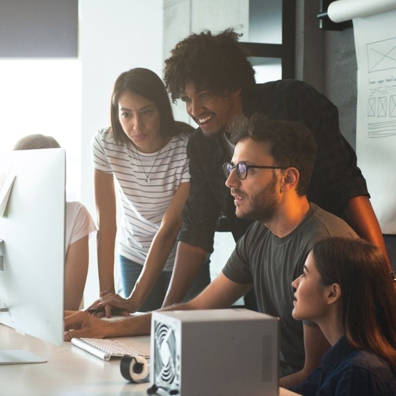 Four people gathered around a computer monitor in an office, collaborating on their project communication plan. Papers and a whiteboard with diagrams are visible in the background as they focus intently on the screen.