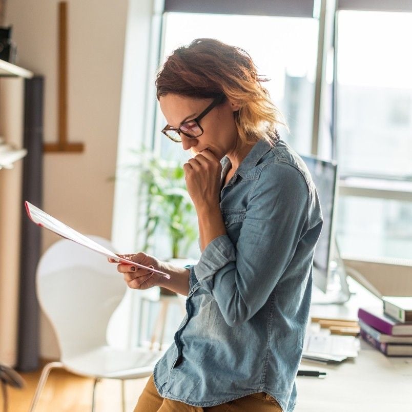 A person wearing glasses and a denim shirt sits on a desk, looking thoughtfully at documents in hand—perhaps handling contract management—with one hand near their mouth. A computer, chair, and bright window are visible in the background.
