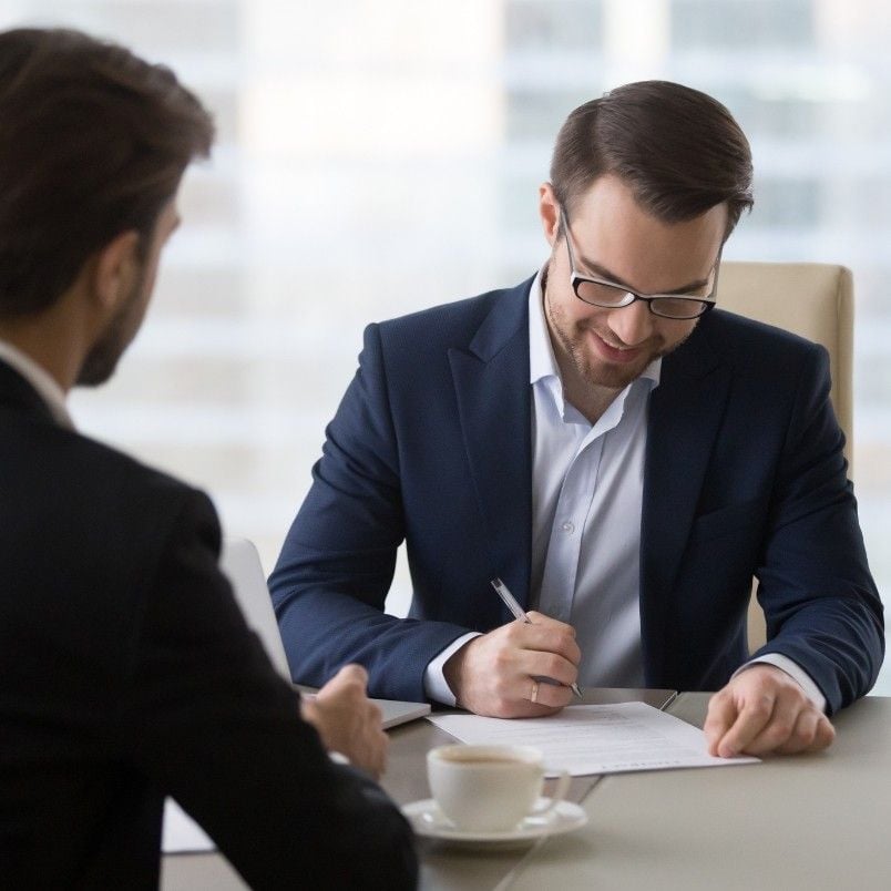 Two men in business suits sit at a table. One man smiles as he signs documents with a pen, while the other listens, illustrating smooth contract management in a bright office setting. A cup of coffee rests on the table between them.