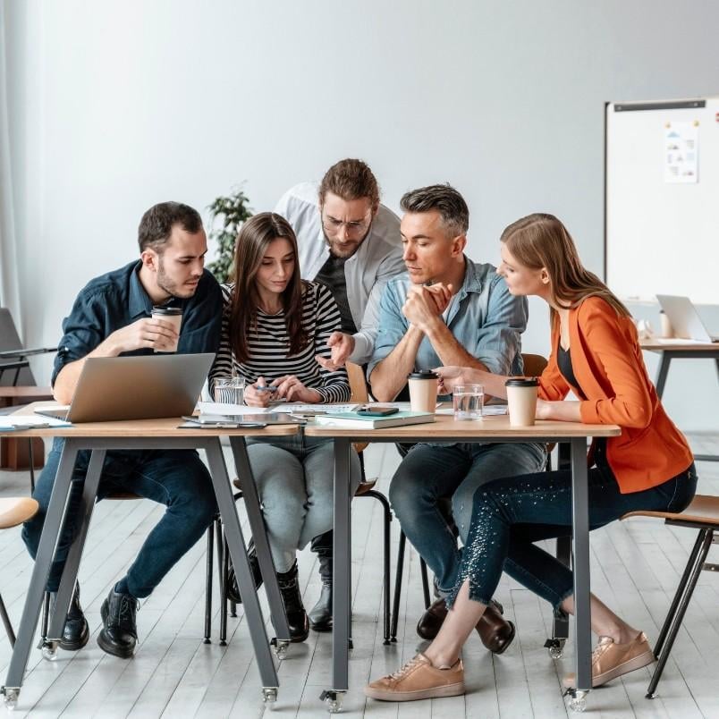 Five people sit around a table in a modern office, collaborating on documents, a laptop, and a wireframe template. They appear focused and engaged, with coffee cups and papers scattered across the table.