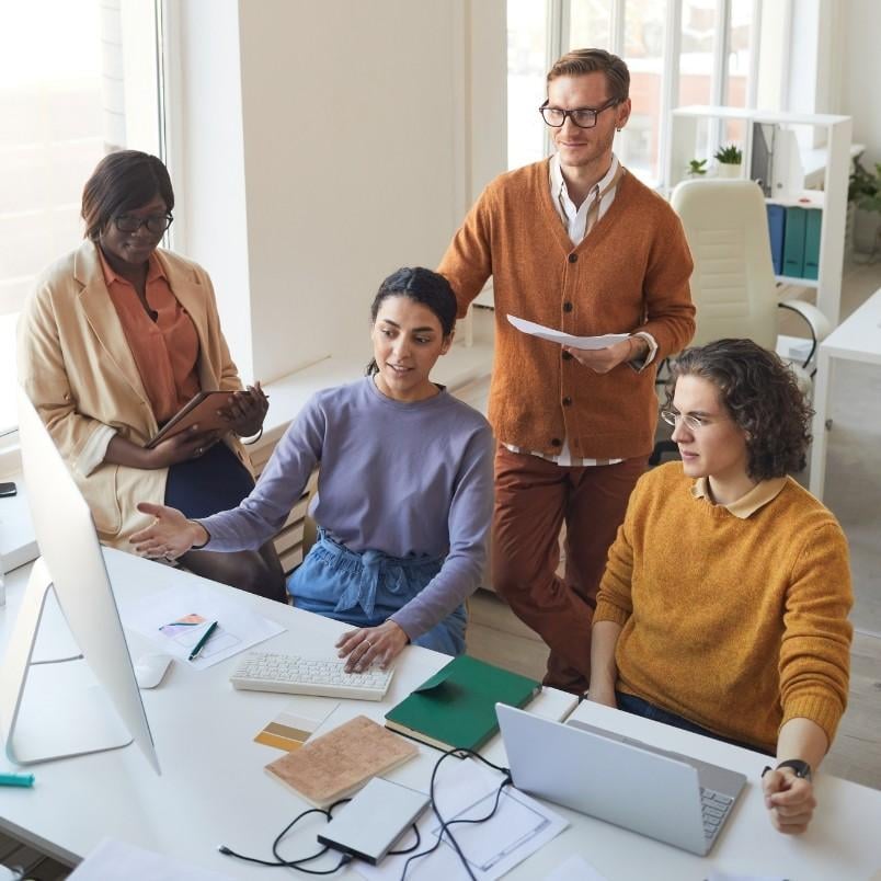 Four people in casual clothes gather around a desk in a bright office, looking at a computer monitor. One person gestures toward the screen, sparking a collaborative discussion about the wireframe template displayed.