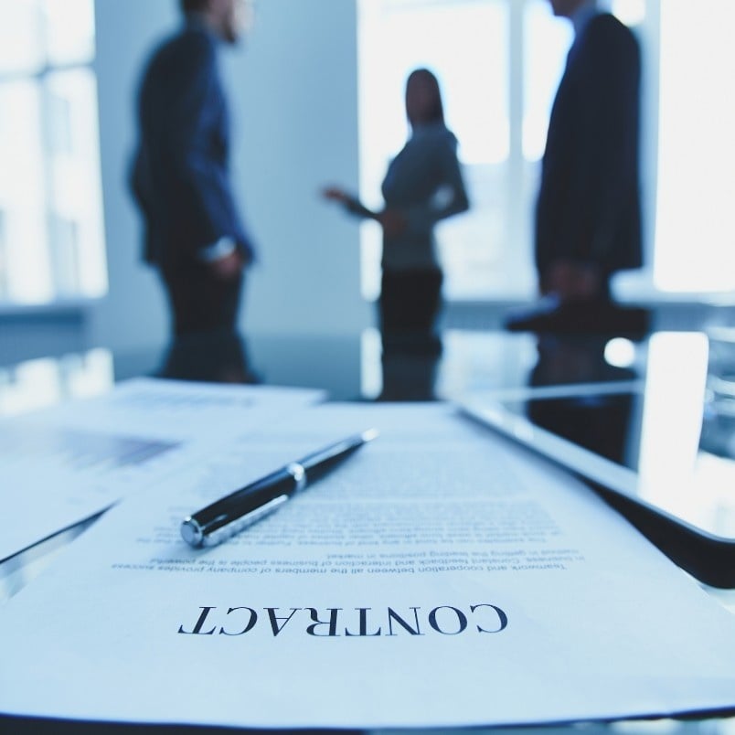 A contract and pen are on a glass table in focus, symbolizing contract management, while three people stand and talk in the blurred background in a bright office setting.
