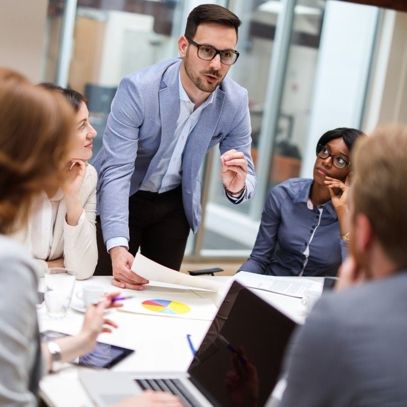 A man in a light blue blazer demonstrates agile skills as he stands and speaks to colleagues during a meeting, holding papers with a colorful pie chart, while others sit around a table with laptops and documents.