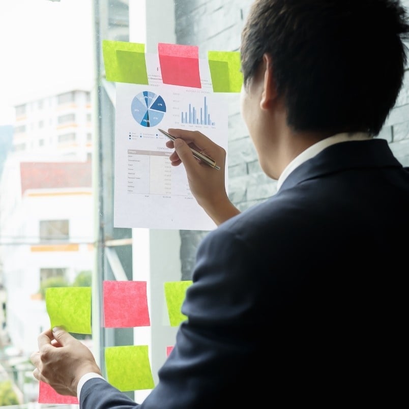 A man in a suit analyzes charts and graphs on a paper taped to a glass wall, surrounded by colorful sticky notes—a scene illustrating the project manager role in scrum as he reviews data near a window, pen in hand.