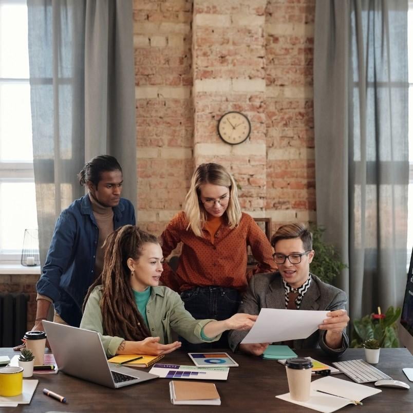 Four people gather around a table covered with laptops, notebooks, and coffee cups, discussing project management and business process improvements in a modern office with brick walls and large windows.