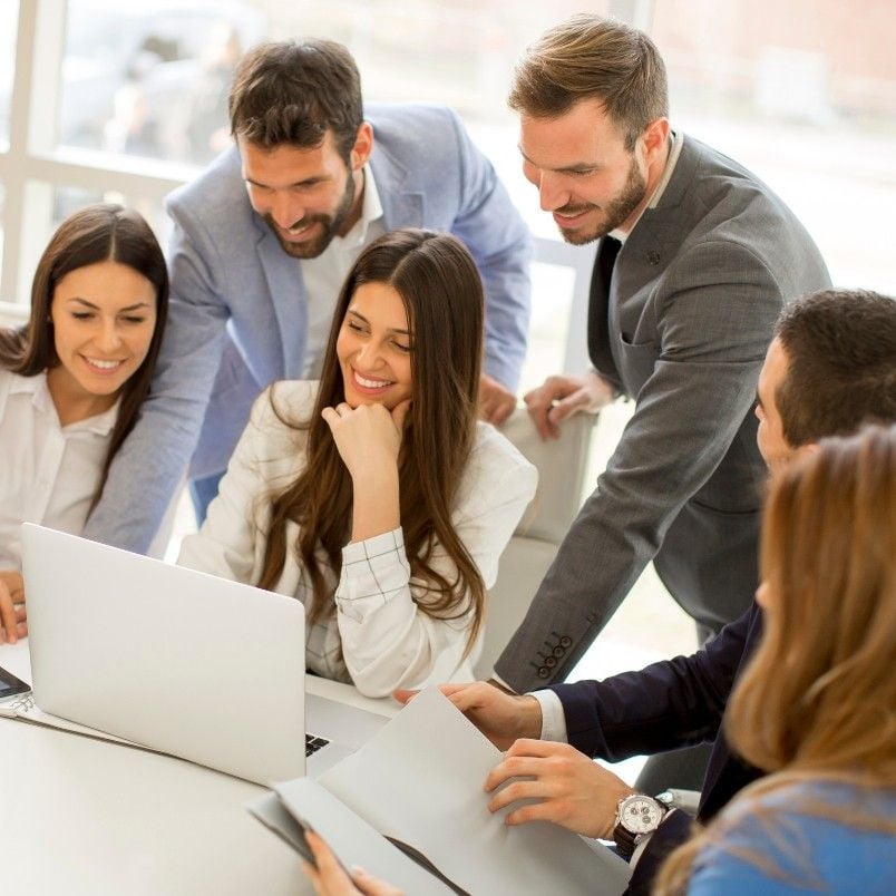 Five people in business attire gather around a laptop, smiling and discussing their project communication plan in a bright office setting, with papers and documents on the table.