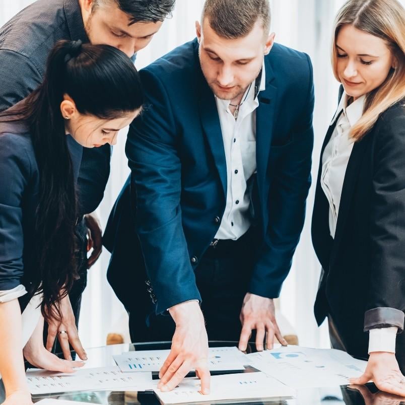 Four business professionals in formal attire gather around a table, closely examining papers and charts. One man points at a document, while the others look on attentively, discussing their project communication plan.