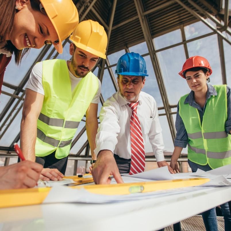 Four construction workers, wearing hard hats and safety vests, gather around a table with blueprints and tools, discussing Construction Project Management plans inside a modern building with a glass roof.